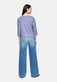 Lila Bluse mit drei Viertelärmeln, kombiniert mit weit geschnittenen, hellblauen Jeans, die ein allmähliches Nietenmuster in der Nähe des Saums aufweisen.