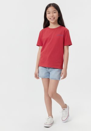 Fată zâmbitoare cu păr lung și închis la culoare, purtând o cămașă roșie cu mâneci scurte, pantaloni scurți din denim albastru deschis și adidași albi, stând pe un fundal simplu.