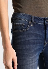Tumma sininen farkkutakki, jossa on kevyt haalistuminen, viisi taskua ja näkyvät ompeluyksityiskohdat. Slim fit -malli, suora lahje.