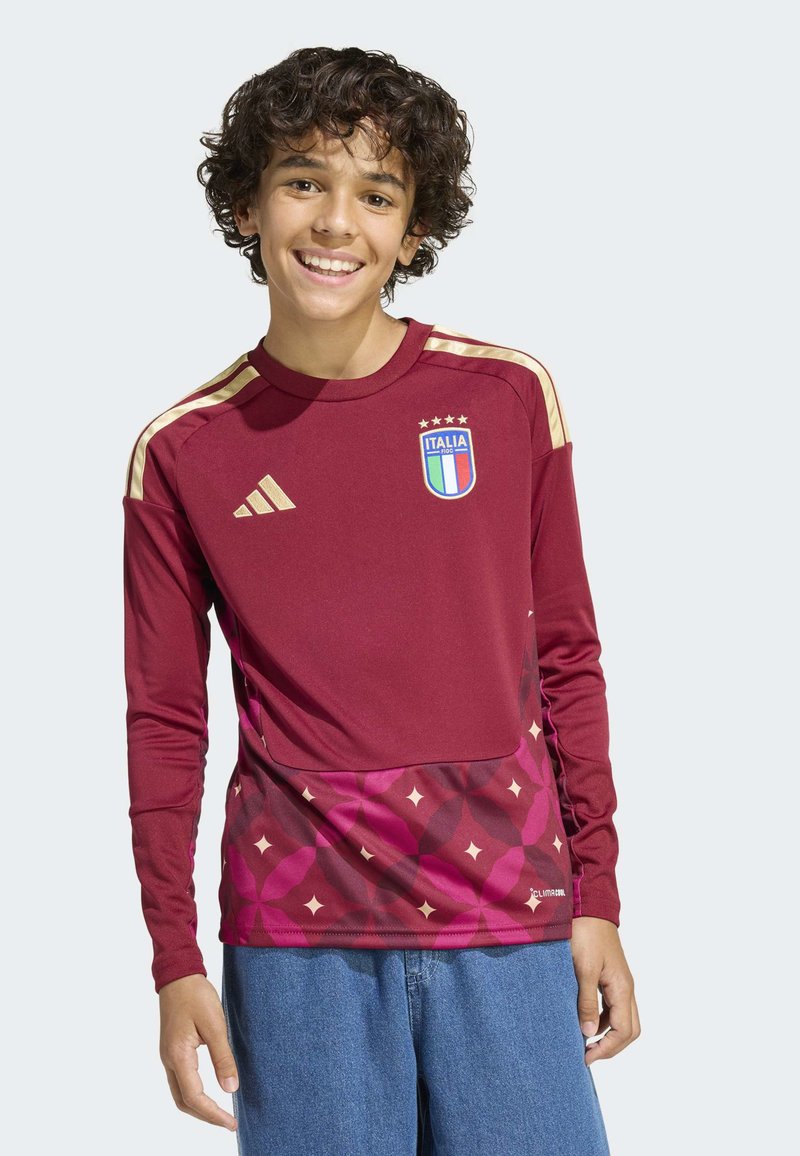 Smiling teenage boy with curly hair wearing a maroon Italian football jersey with gold stripes and blue jeans, standing against a light background.