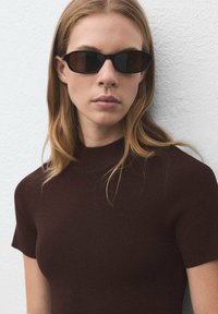 Des lunettes de soleil noires rectangulaires avec des verres sombres portées avec un haut à col montant côtelé marron, sur un fond blanc uni.