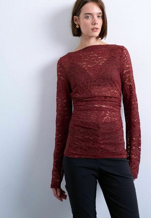 Kvinde iført en tætsiddende, langærmet bordeaux blomstret blondebluse og sorte bukser, stående foran en ensfarvet lysegrå væg.