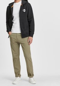 Sudadera negra con cremallera y logo blanco, con acentos naranjas en el interior de la capucha; combinada con chinos color caqui y zapatillas blancas.