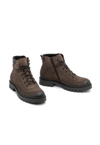 Botas de tobillo de ante marrón con cordones en la parte delantera, cremallera lateral y suelas de goma. Presentan acentos metálicos y un collar acolchado.