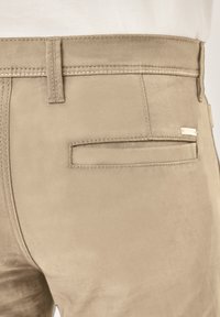 Beige Wildlederhose mit glatter Textur, ausgestattet mit einer Gesäßtasche und Kontrastnähten. Ein Metall-Logoschild ist am Bund sichtbar.