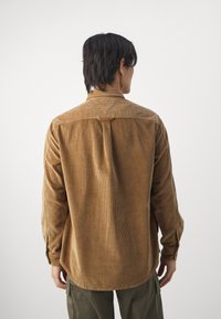 Brown corduroy shirt with a relaxed fit, vertical ribbed texture, long sleeves, and a back yoke detail. Tailored collar and rounded hem.