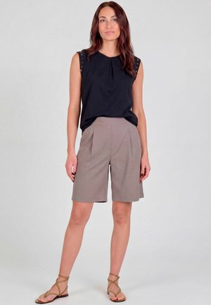 Ung kvinde med langt brunt hår iført en sort ærmeløs top, taupe knængående shorts og tan farvede sandaler med stropper, står foran en ensfarvet baggrund.