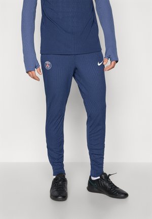 Mann trägt blaue, gerippte Paris Saint-Germain Nike Trainingshose und passendes Oberteil mit Daumenlöchern sowie schwarze Nike-Sneaker mit weißen Socken.