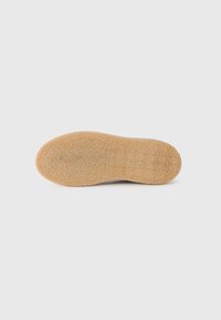 Semelle d'une chaussure, en matériau en caoutchouc texturé de couleur brun clair avec une finition lisse et un détail de logo en relief.