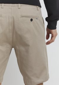 Beige, strukturerede shorts med en baglomme med knaplukning. Falde ved kanten og en flad talje kan ses.