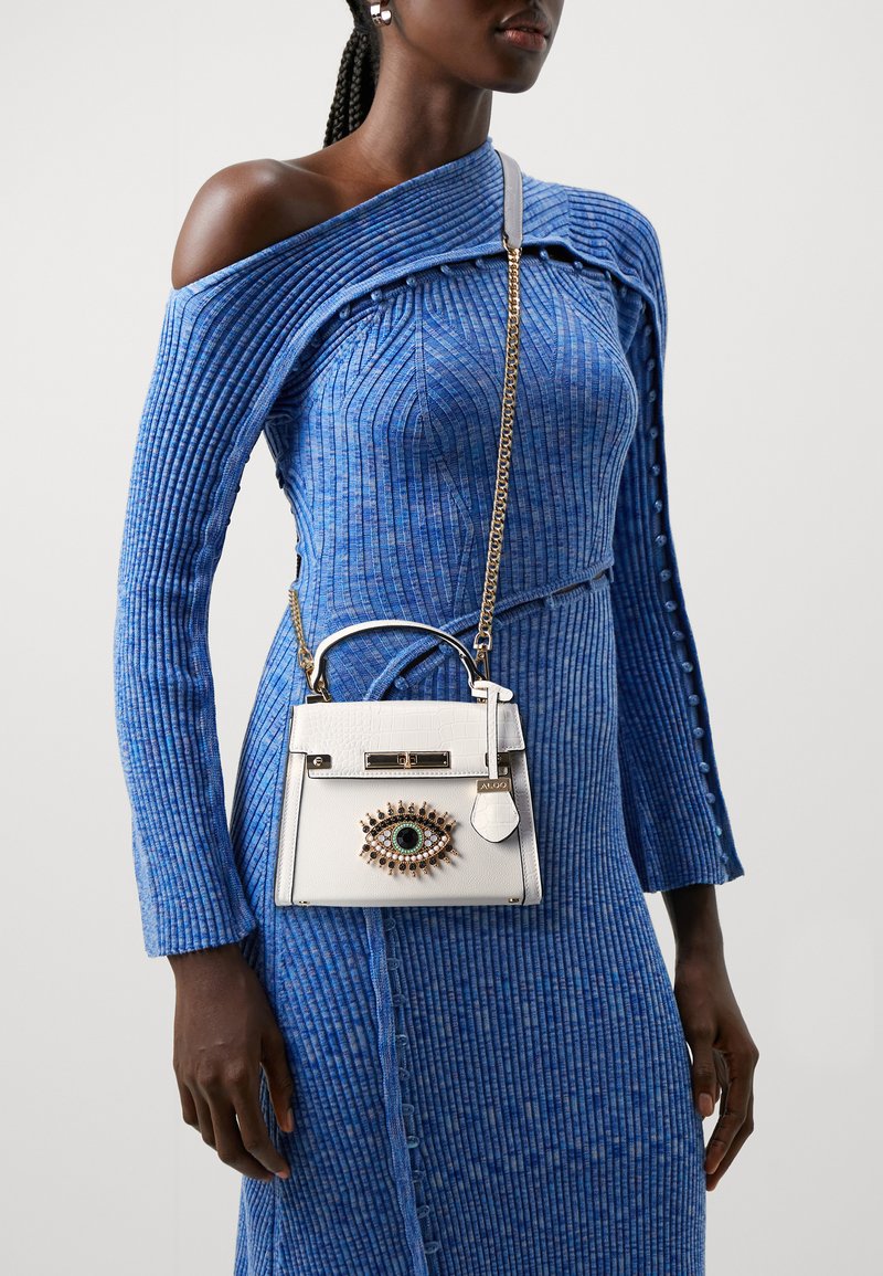 White leather handbag with crocodile texture, gold chain strap, decorated with an eye motif featuring an emerald centerpiece, paired with a blue ribbed dress.