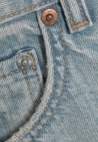 Gros plan sur un jean en denim bleu clair montrant des coutures détaillées et des rivets métalliques sur les poches et la ceinture.