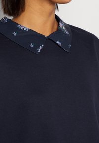 Námořnická mikina s květinově vzorovaným modrým límcem. Měkká textura, uvolněný střih a klasický design viditelný v detailech límce.