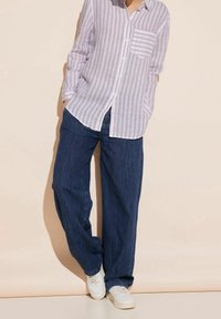 Camicia a righe lilla chiaro e bianca con taschino sul petto, abbinata a pantaloni larghi in denim scuro e sneaker bianche.