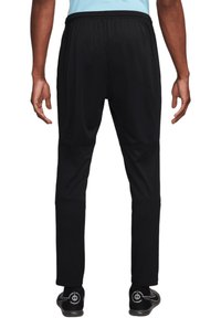 Pantalons de sport noirs avec une taille élastique, coupe ajustée et panneaux texturés aux genoux, associés à des chaussures de sport noires avec des accents gris.