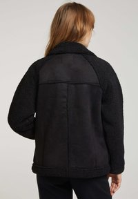Veste noire avec un tissu texturé et flou sur les manches et le dos. Partie centrale lisse, col arrondi et détails cousus sur les coutures.