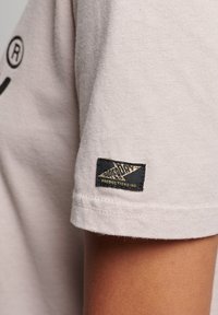Světle béžové bavlněné tričko s krátkými rukávy, které má na rukávu černou nášivku s trojúhelníkovým designem a bílým textem pro branding.