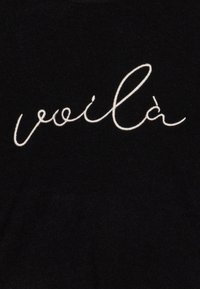 Fekete szövet, amelyen fehér hímzett szöveg található, amely a "voilà" szót tartalmazza. Egyszerű, sima textúra, laza dizájn.