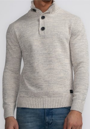 Man wearing light gray knitted sweater with two black buttons on high collar, paired with blue jeans, against plain background.