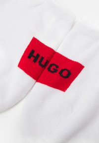 Balti medvilniniai kojinės su raudona stačiakampio formos juosta, ant kurios juodu tekstu užrašyta "HUGO". Kojinės pasižymi lygiu audiniu ir paprastu dizainu.