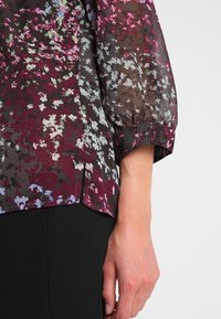 Blouse transparente à motif floral dans des tons foncés de violet, rouge et blanc. Présente une coupe décontractée et des manches trois-quarts bouffantes.