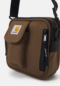 Borsa a spalla in tela marrone con doppia cerniera, tasca frontale, tracolla regolabile e una patch del logo Carhartt gialla sulla parte anteriore.