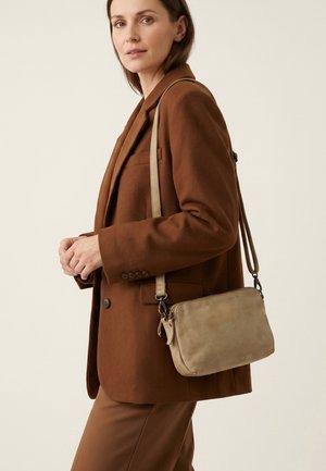 Femme portant un costume marron avec un sac bandoulière en cuir beige clair sur un fond clair uni.