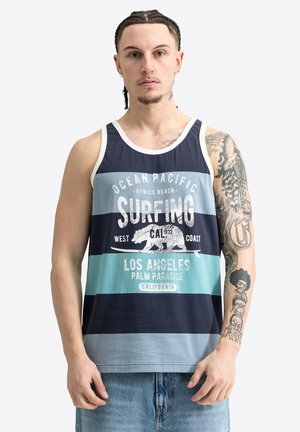 Mężczyzna z warkoczami ubrany w pasiasty tank top z napisami surfing i Los Angeles, pokazujący wytatuowaną prawą rękę i szorty jeansowe.