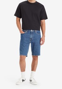Man som bär en svart t-shirt, midilånga blå denimshorts, vita strumpor och vita sneakers med svarta detaljer, stående mot vit bakgrund.