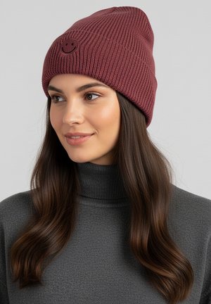 Maroon knit beanie with a ribbed texture and a small black smiley face patch on the front. Worn with a gray high-neck sweater.