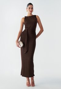 Een bruine, mouwloze maxi-jurk met een drapering aan de zijkant in een knoopontwerp. Glad van textuur, aansluitende silhouet, gecombineerd met een witte clutch en sandalen met banden.