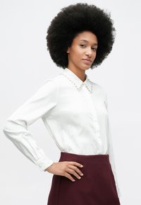 Woman with afro hairstyle wearing white blouse with pearl collar and burgundy skirt, standing with hand on hip against white background.