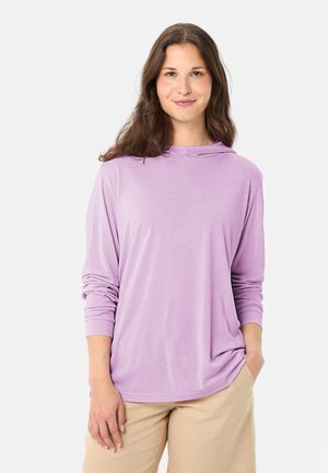 Vrouw met lang bruin haar, gekleed in een loszittend lavendelkleurig shirt met lange mouwen en beige broek, staand met één hand in haar zak tegen een effen achtergrond.