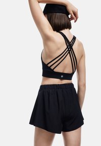 Mujer vista de espaldas, vistiendo un sujetador deportivo negro con tirantes, pantalones cortos negros y una gorra negra, con un brazo levantado detrás de su cabeza.