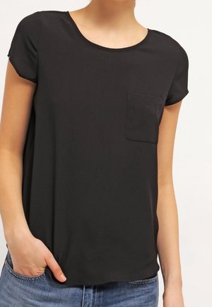 Kvinde iført en ensfarvet sort T-shirt med korte ærmer og brystlomme, kombineret med blå jeans, stående med den ene hånd i lommen.