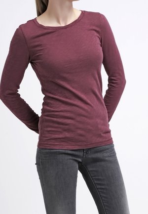 Mujer con una camiseta ajustada de manga larga color granate y vaqueros gris oscuro, de pie con las manos detrás de la espalda contra un fondo liso.