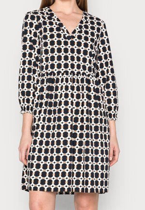 Frau trägt ein knielanges Kleid mit schwarz-weißem geometrischem Kreis-Muster und Dreiviertelärmeln vor einfarbigem Hintergrund.