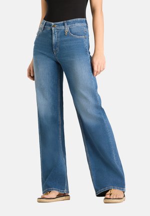 Vrouw draagt hoog getailleerde wijde blauwe jeans met voorzakken en een knoop, gecombineerd met een zwarte top en beige sandalen.