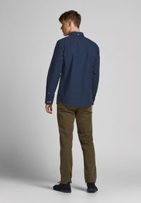 Camisa de manga larga azul marino con cuello de botón, con pliegue en la parte posterior, combinada con pantalones marrón claro y zapatos oscuros. Vista trasera, fondo liso.