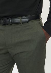 Mörkgröna byxor i texturerad tyg, med ett svart läderbälte med rektangulär spänne och sidofickdetaljer.