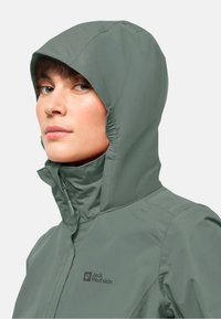 Veste imperméable verte avec capuche ajustée, col haut et accents élastiques. Présente un logo sur la poitrine et une texture lisse et mate.