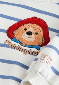 Sweat-shirt blanc à rayures bleues, avec un ours Paddington brodé en relief et une étiquette indiquant : « Veuillez prendre soin de cet ours. Merci. »