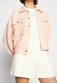 Femme portant une veste pied-de-poule rose et blanche sur un t-shirt blanc et une jupe texturée crème sur un fond uni.