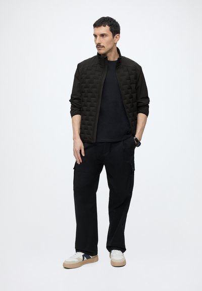 Homme portant une veste matelassée noire, un pantalon noir et des baskets blanches à semelles beiges, debout avec une main dans la poche.