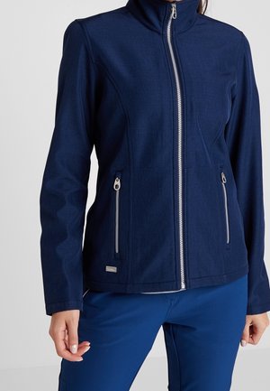 Femme portant une veste zippée bleu marine avec deux poches latérales zippées et un pantalon bleu marine assorti sur un fond clair uni.