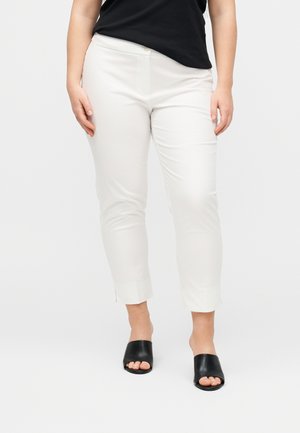 Γυναίκα που φοράει λευκό στενό παντελόνι μέχρι τον αστράγαλο, μαύρη μπλούζα με κοντό μανίκι και μαύρα mule παπούτσια με ανοιχτά δάχτυλα, στέκεται σε απλό ανοιχτό φόντο.