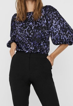 Vrouw die een paars-zwart patroon blouse draagt met pofmouwen en een zwarte high-waisted broek, handen in de zakken.