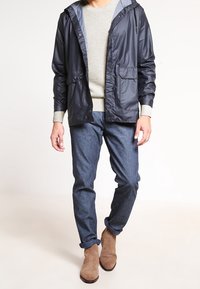 Coupe-vent léger bleu marine avec capuche, deux poches frontales, assorti à un pull gris, un jean en denim foncé et des bottines en suède marron.