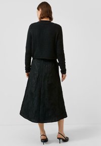 Schwarzer Pullover mit strukturiertem Strick, kombiniert mit einem bodenlangen schwarzen Rock mit floralen Mustern. Die Schuhe sind schwarz mit einem niedrigen Absatz.