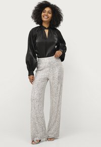 Blusa nera di satin con scollo a chiave. Pantaloni larghi in paillettes argentate con texture a righe verticali. Modella in piedi su uno sfondo neutro.